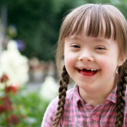 Criança com síndrome de Down sorri para a foto. É uma menina branca, com cabelos castanhos claros divididos em duas tranças e uma franja. Ela veste uma camisa quadriculada na cor rosa. Ao fundo, desfocado, está um jardim.