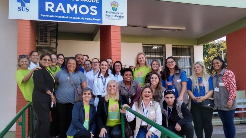 Visita da OPAS PIM Rubem Berta. A foto mostra um grupo grande de profissionais de saúde em frente a Unidade Básica de Saúde. 
